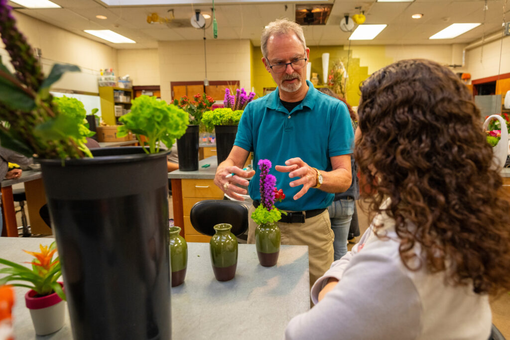 Instructor with floral arrangement in front of him, talking to a student.